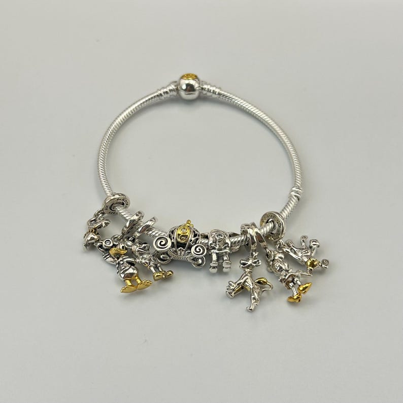 May include: Silver charm bracelet with multiple charms. Charms include a carriage, characters with gold accents, and other decorative elements. The bracelet is on a white background.