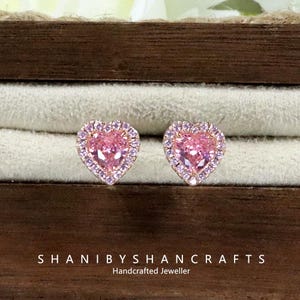 Könnte beinhalten: Ein Paar herzförmige Ohrstecker mit rosa Mittelstücken und einem Kranz aus kleineren, hellvioletten Steinen. Die Ohrringe sind in einem goldfarbenen Metall gefasst. Der Text "SHANIBYSHANCRAFTS Handcrafted Jeweller" ist sichtbar.