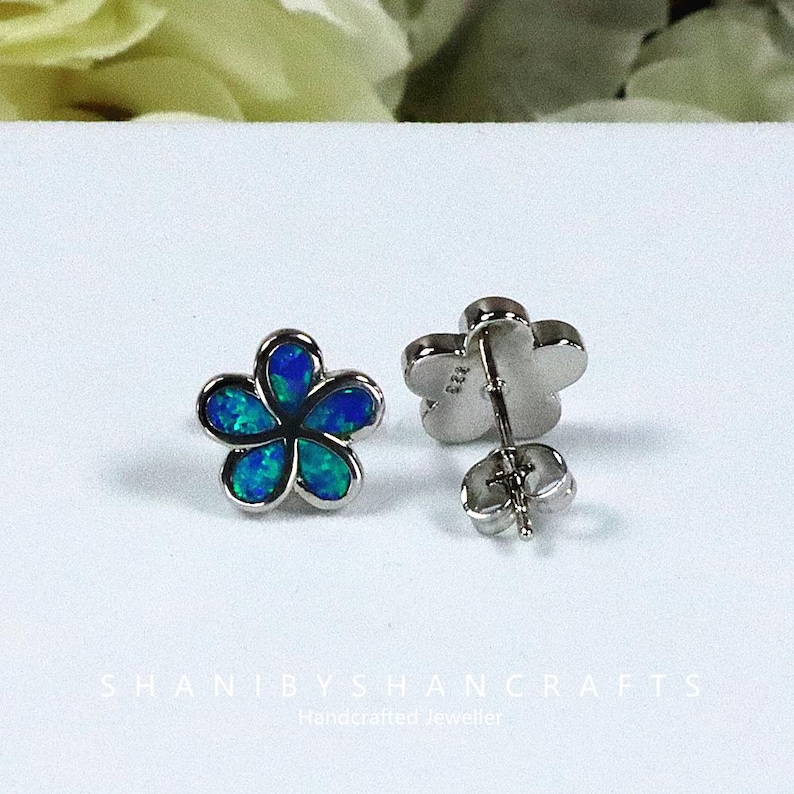 May include: A pair of flower-shaped stud earrings. Each earring features a five-petal design with a silver-colored border and a blue-green opal center. The earrings are displayed on a white surface. The text "SHANIBYSHANCRAFTS" is visible.