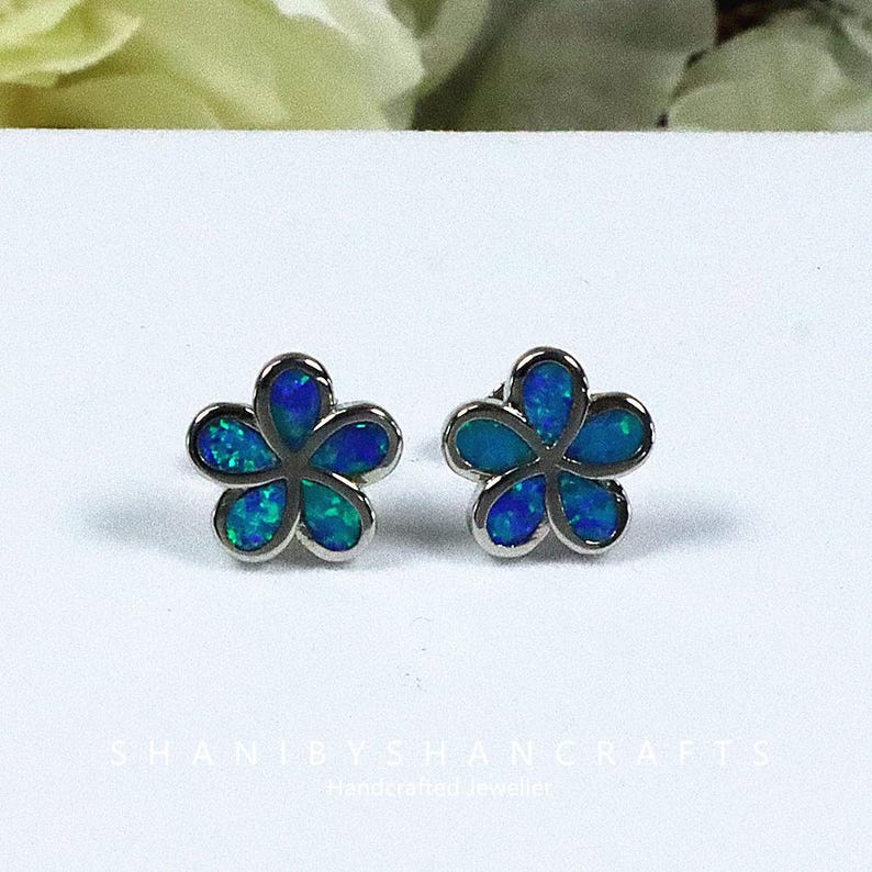 May include: A pair of flower-shaped stud earrings. Each earring features five petals with a silver-colored border. The petals are filled with a blue and green iridescent material. The earrings are displayed on a white surface.