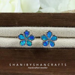 May include: A pair of flower-shaped stud earrings with a silver-colored setting. The petals are filled with blue and turquoise-colored opal. The earrings are displayed in a wooden jewelry box. The text "SHANIBYSHANCRAFTS Handcrafted Jeweller" is visible.