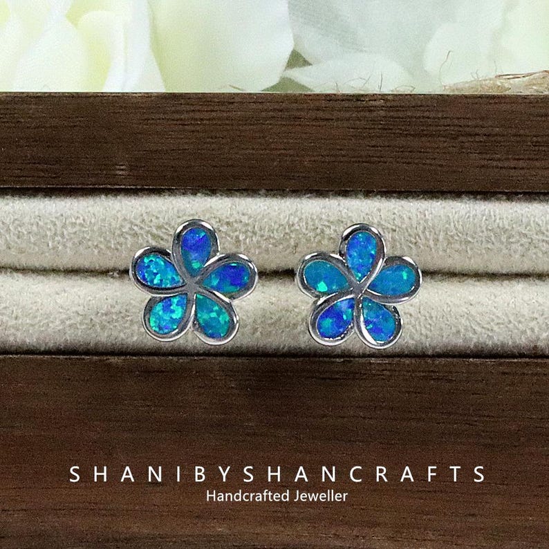 May include: A pair of flower-shaped stud earrings with a silver-colored setting. The petals are filled with blue and turquoise-colored opal. The earrings are displayed in a wooden jewelry box. The text "SHANIBYSHANCRAFTS Handcrafted Jeweller" is visible.