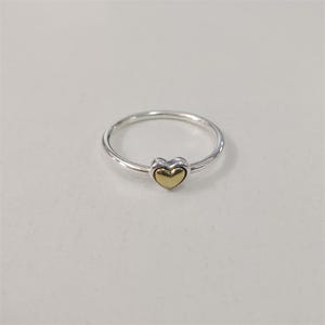 May include: A silver ring with a gold heart-shaped center. The ring is simple and elegant, with a thin band and a small heart. The heart is a bright gold color, and the band is a shiny silver.