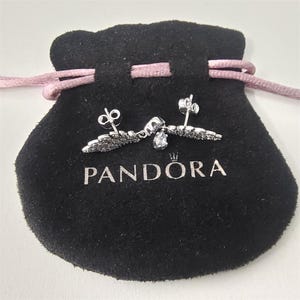 K&ouml;nnte beinhalten: Silberne Ohrringe mit Fl&uuml;geldesign, schwarzen Akzenten und einem tropfenf&ouml;rmigen Edelstein, pr&auml;sentiert auf einem schwarzen Samtbeutel mit dem Markennamen "PANDORA" in silberner Schrift. Der Beutel hat einen rosa Kordelzug.