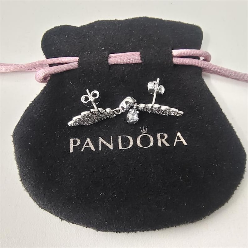 K&ouml;nnte beinhalten: Silberne Ohrringe mit Fl&uuml;geldesign, schwarzen Akzenten und einem tropfenf&ouml;rmigen Edelstein, pr&auml;sentiert auf einem schwarzen Samtbeutel mit dem Markennamen "PANDORA" in silberner Schrift. Der Beutel hat einen rosa Kordelzug.