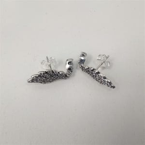 K&ouml;nnte beinhalten: Ein Paar silberfarbene Ohrstecker. Jeder Ohrring hat ein fl&uuml;gelf&ouml;rmiges Design, besetzt mit kleinen, klaren Edelsteinen. Die Ohrringe werden auf einer wei&szlig;en Oberfl&auml;che pr&auml;sentiert.