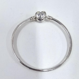 May include: A silver bangle bracelet with a heart-shaped clasp. The clasp has the letter "O" in the centre. The bracelet is made of a smooth, shiny metal and is designed to be worn on the wrist.