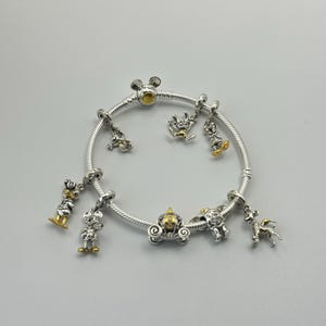 May include: Silver charm bracelet with gold and silver charms. The charms include a carriage, a deer, and several cartoon character figures. The bracelet is on a white background.