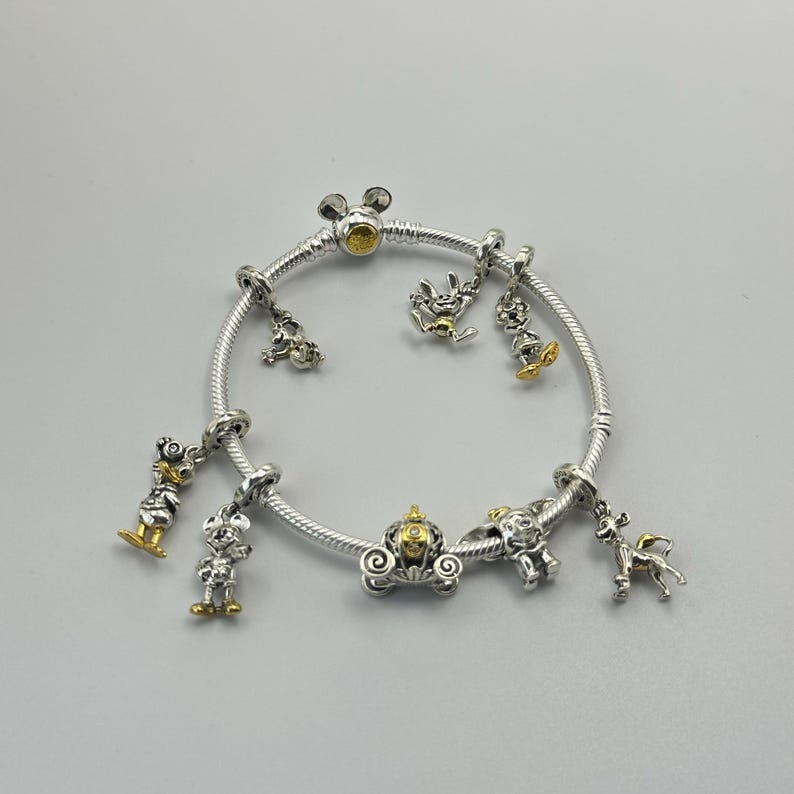 May include: Silver charm bracelet with gold and silver charms. The charms include a carriage, a deer, and several cartoon character figures. The bracelet is on a white background.
