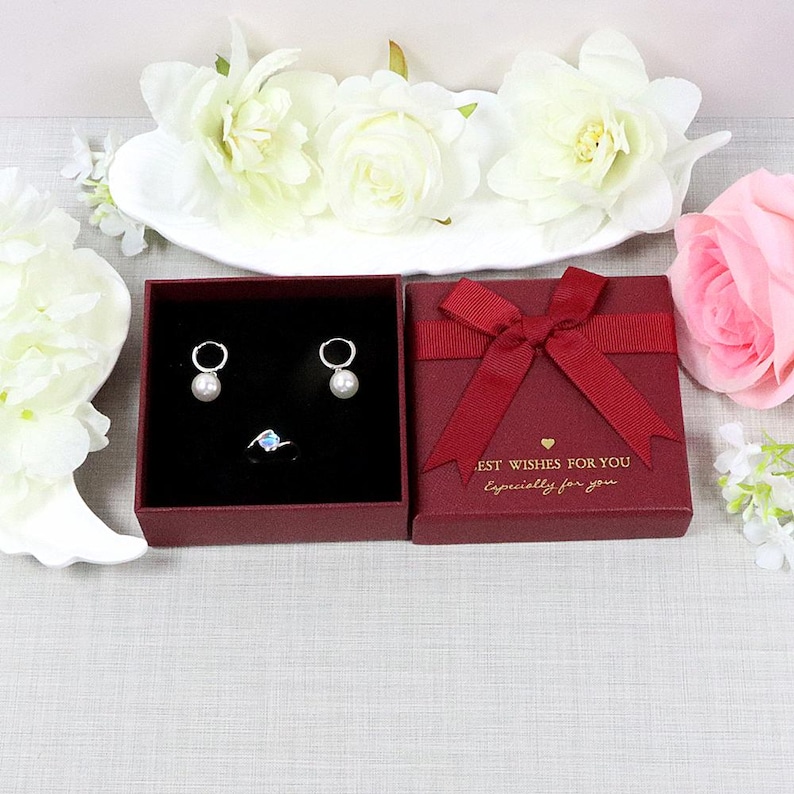 May include: A jewelry set featuring a pair of pearl earrings and a ring with a blue gemstone, presented in a red gift box with a bow. The box has the text "BEST WISHES FOR YOU Especially for you."