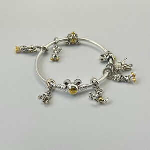 May include: A silver charm bracelet with gold accents and various Disney-themed charms. Charms include a Mickey Mouse head, a pumpkin carriage, and other character figures. The bracelet is displayed on a light gray surface.