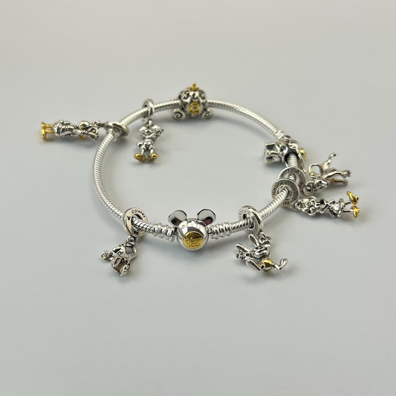 May include: A silver charm bracelet with gold accents and various Disney-themed charms. Charms include a Mickey Mouse head, a pumpkin carriage, and other character figures. The bracelet is displayed on a light gray surface.