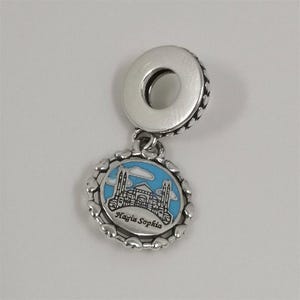 Può includere: Un charm in argento con un ciondolo rotondo raffigurante un edificio con minareti su sfondo di cielo blu. Il testo "Hagia Sophia" è in basso. Il charm ha un bordo a forma di cuore.