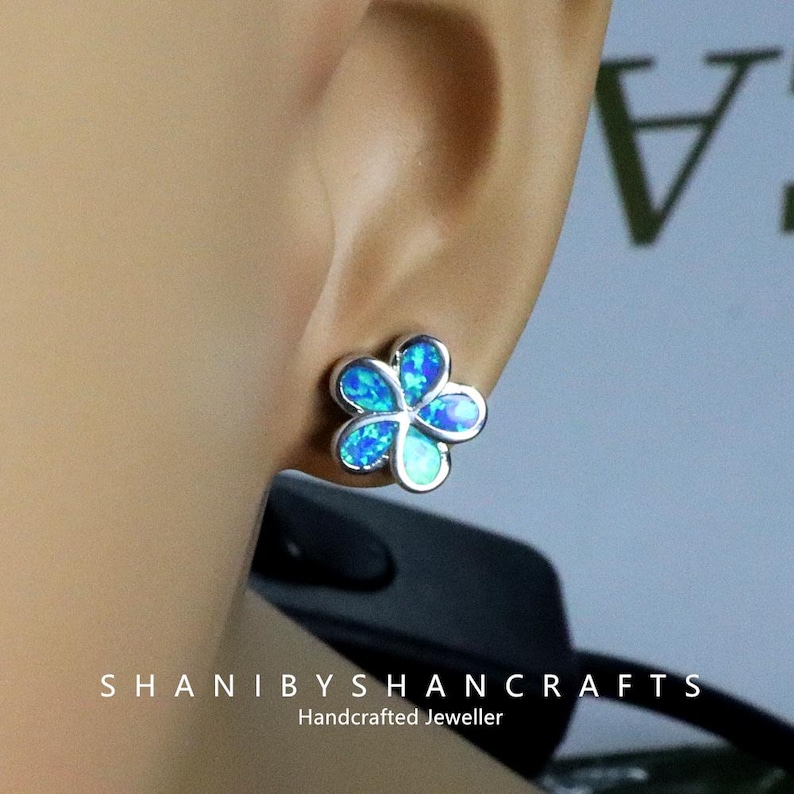 May include: A silver-toned flower stud earring with blue opal petals. The earring is worn on an ear. The image also shows the text "SHANIBYSHANCRAFTS" and "Handcrafted Jeweller".