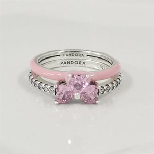 Op de afbeelding: Drie zilveren ringen. Eén ring heeft een lichtroze emaille band. Een andere ring heeft een rij kleine, heldere stenen. De derde ring heeft drie roze hartvormige stenen. Het woord "PANDORA" is op de ringen gegraveerd.