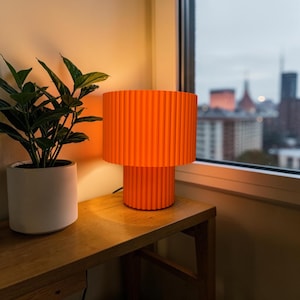 Może przedstawiać: Pomarańczowa lampa stołowa z plisowanym kloszem i cylindryczną podstawą. Lampa stoi na drewnianym stole obok rośliny doniczkowej. W tle widać panoramę miasta przez okno.