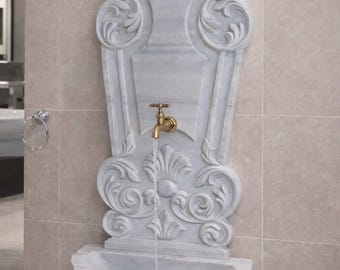 May include: A marble wall fountain with ornate carvings and a brass faucet. The fountain features a basin and a decorative back panel with scrollwork and a shell-like top. Water flows from the faucet into the basin. The fountain is mounted on a tiled wall.
