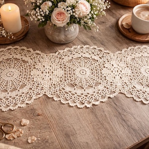 Könnte beinhalten: Cremefarbener, handgefertigter Häkel-Tischläufer mit detailliertem kreisförmigem Muster. Der Läufer wird auf einer Holzoberfläche zusammen mit einer Kerze, Blumen und einer Tasse Kaffee präsentiert. Ein dekoratives Stück für die Inneneinrichtung.