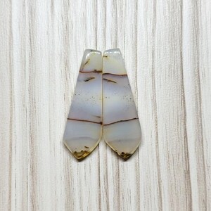 May include: Two polished, elongated gemstone cabochons. The stones are primarily white with bands of brown and gray, and small dark spots. The cabochons have a teardrop shape with a flat back and a polished, slightly rounded top. The stones are set against a light wood grain background.