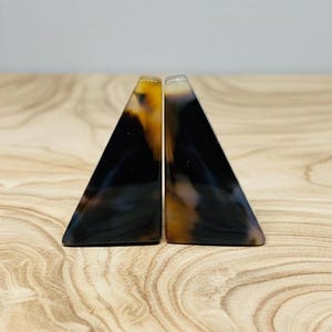 Pode incluir: Dois suportes de livros triangulares com um padrão de tartaruga. Os suportes de livros são feitos de um material brilhante, com um gradiente de cores do castanho escuro ao âmbar. Estão colocados numa superfície de madeira clara.