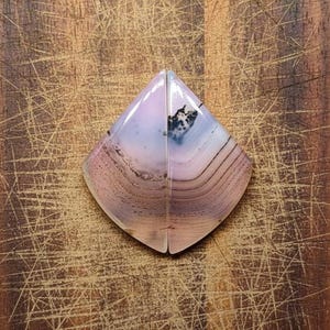 Puede incluir: Dos mitades de piedras preciosas pulidas a juego. Las piedras tienen forma de diamante, con un degradado de color de rosa pálido a lavanda y blanco. Bandas e inclusiones de color marrón oscuro y negro añaden detalles. Las piedras se exhiben sobre una superficie de madera.