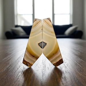 Könnte beinhalten: Zwei Buchstützen aus Achat, jede mit einem einzigartigen Muster aus beigen, braunen und weißen Bändern. Die Buchstützen sind so geschnitten, dass sie eine dreieckige Form bilden, mit einer dunkelgrauen Mitte. Sie werden auf einem Holztisch präsentiert.