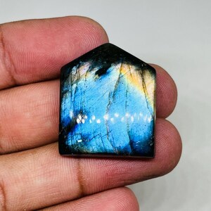 May include: A house-shaped labradorite gemstone featuring a polished surface with iridescent flashes of electric blue, deep navy, and golden yellow. The stone displays natural internal fractures and a smooth, flat face with a pointed top edge.