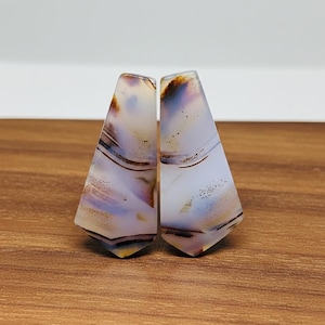 May include: Two polished agate stones with a triangular shape. The stones feature a mix of colors, including white, brown, and purple, with intricate banding patterns. The stones are displayed on a wooden surface.