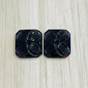 May include: Two dark purple, rectangular gemstones with carved floral designs. The gemstones have a polished finish and are set against a light wood grain background. The carved details create a three-dimensional effect.