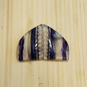 May include: Two matching, polished, semi-circular stones with a carved central design. The stones feature alternating bands of purple, clear, and beige hues. The stones are displayed on a light brown, wooden surface.
