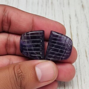 May include: Two polished, dark purple gemstone cabochons with a grid-like carved pattern. The stones are held in a hand, showcasing their color and texture. The cabochons have a rounded shape with a flat back.