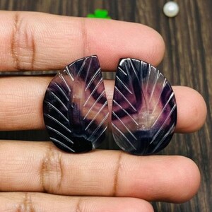 May include: Two carved purple and white gemstone pendants. Each pendant has a teardrop shape with a leaf-like design carved into the surface. The gemstones are held in a person's hand.