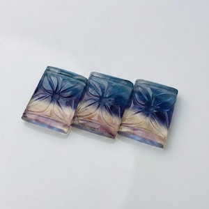 May include: Three rectangular, carved fluorite stones. Each stone features a floral design with a gradient of colors, transitioning from blue to purple and cream. The stones are arranged on a white surface.