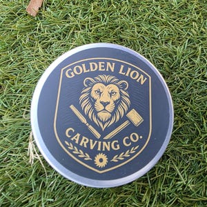 May include: A round, black and silver container with the text "GOLDEN LION CARVING CO." in gold lettering. The container features a lion's head emblem with carving tools. The container is sitting on green grass.