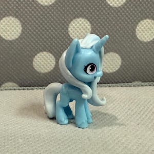 Könnte beinhalten: Ein kleines, hellblaues Spielzeugpferd mit weißer Mähne und Schweif. Das Pony hat ein einzelnes Horn und große Augen mit rosa Akzenten. Das Spielzeug steht auf einer weißen Oberfläche mit grauem Polka-Dot-Hintergrund.