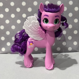 Könnte beinhalten: Ein rosa Spielzeugpferd mit lila Haaren und einem weißen Flügel an der Seite. Das Pony hat grüne Augen und ein Symbol an der Flanke. Das Pony steht auf einer grauen Oberfläche mit weißen Punkten.