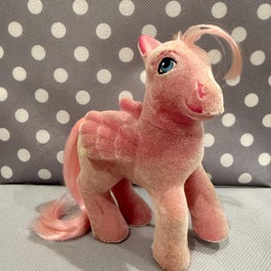 Könnte beinhalten: Ein Vintage-Spielzeugpferd in Rosa mit Flügeln, einem langen rosa Schweif und einer rosa Mähne. Das Pony hat blaue Augen und steht auf allen vier Beinen. Das Spielzeug hat eine weiche, samtige Textur und ist ein klassisches Kinderspielzeug.