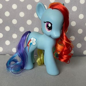 May include: A blue My Little Pony toy with a rainbow mane and tail. The pony has a pink eye and a white cloud with a rainbow and lightning bolt on its flank.