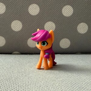 Könnte beinhalten: Eine kleine Ponyfigur in Orange und Lila mit rosa Mähne und Schweif. Das Pony hat türkisfarbene Augen und steht vor einem grauen Hintergrund mit weißen Punkten. Ein Sammlerspielzeug.