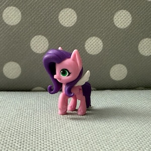 Könnte beinhalten: Eine kleine, rosa und lila Spielzeug-Pony-Figur mit weißen Flügeln. Das Pony hat einen rosa Körper, lila Haare und grüne Augen. Der Hintergrund ist eine graue Oberfläche mit weißen Punkten.