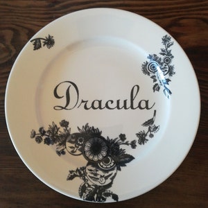 May include: A white ceramic plate with black floral designs and the word "Dracula" written in black cursive script.