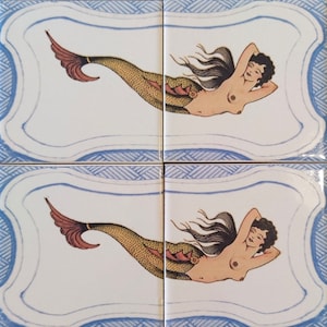 May include: Two ceramic tiles with a blue and white border and a white background. Each tile features a mermaid with long black hair, a red tail, and a yellow and brown fish scale pattern. The mermaid is lying on her side with her head resting on her hand.