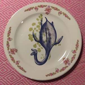 Può includere: Piatto in ceramica bianca con un'illustrazione blu e verde di un plesiosauro. Il piatto ha un bordo floreale con accenti rosa e verdi. Il bordo del piatto è dorato.