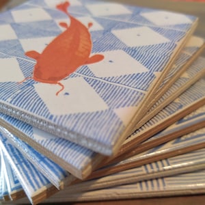 Op de afbeelding: Een stapel keramische onderzetters met een blauw-wit geruit patroon en een oranje visontwerp.