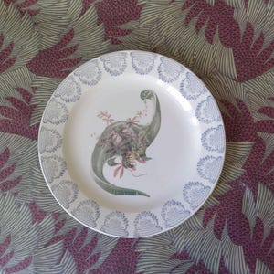 Peut inclure: Assiette en céramique blanche avec une illustration de dinosaure vert et une bordure à motifs bleus. Le dinosaure est orné d'éléments floraux rouges et verts. Un objet décoratif pour la maison.