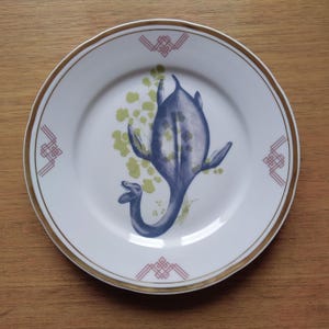 Peut inclure: Une assiette en céramique blanche avec un bord doré et un motif géométrique rose et rouge autour du bord. Le centre de l'assiette présente une illustration bleue et verte d'une créature marine préhistorique.