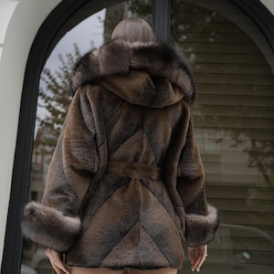 May include: A brown faux fur coat with a hood and a belt. The coat has a chevron pattern and fur trim on the hood and cuffs. The coat is worn by a person standing in front of a window.