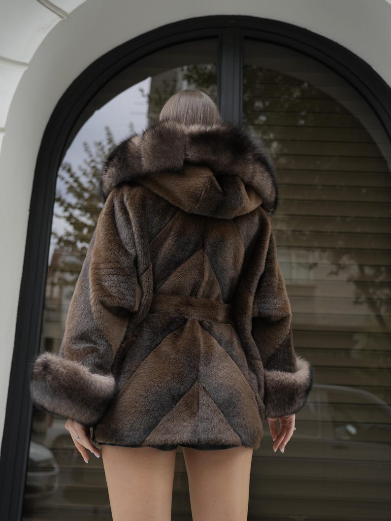 May include: A brown faux fur coat with a hood and a belt. The coat has a chevron pattern and fur trim on the hood and cuffs. The coat is worn by a person standing in front of a window.