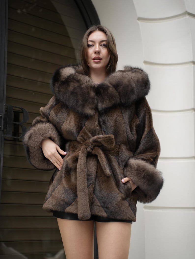 May include: A brown fur coat with a large collar and cuffs. The coat has a tie belt at the waist and is knee-length. The fur appears to be a mix of brown and black tones, creating a textured look. The coat is worn outdoors.