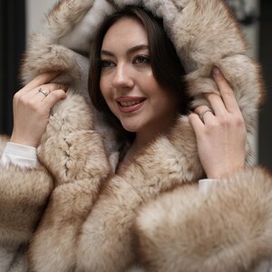 May include: A hooded fur coat with a light brown and cream color scheme. The coat features a large hood and trim around the collar and cuffs. The fur appears soft and luxurious, creating a warm and stylish outerwear piece.